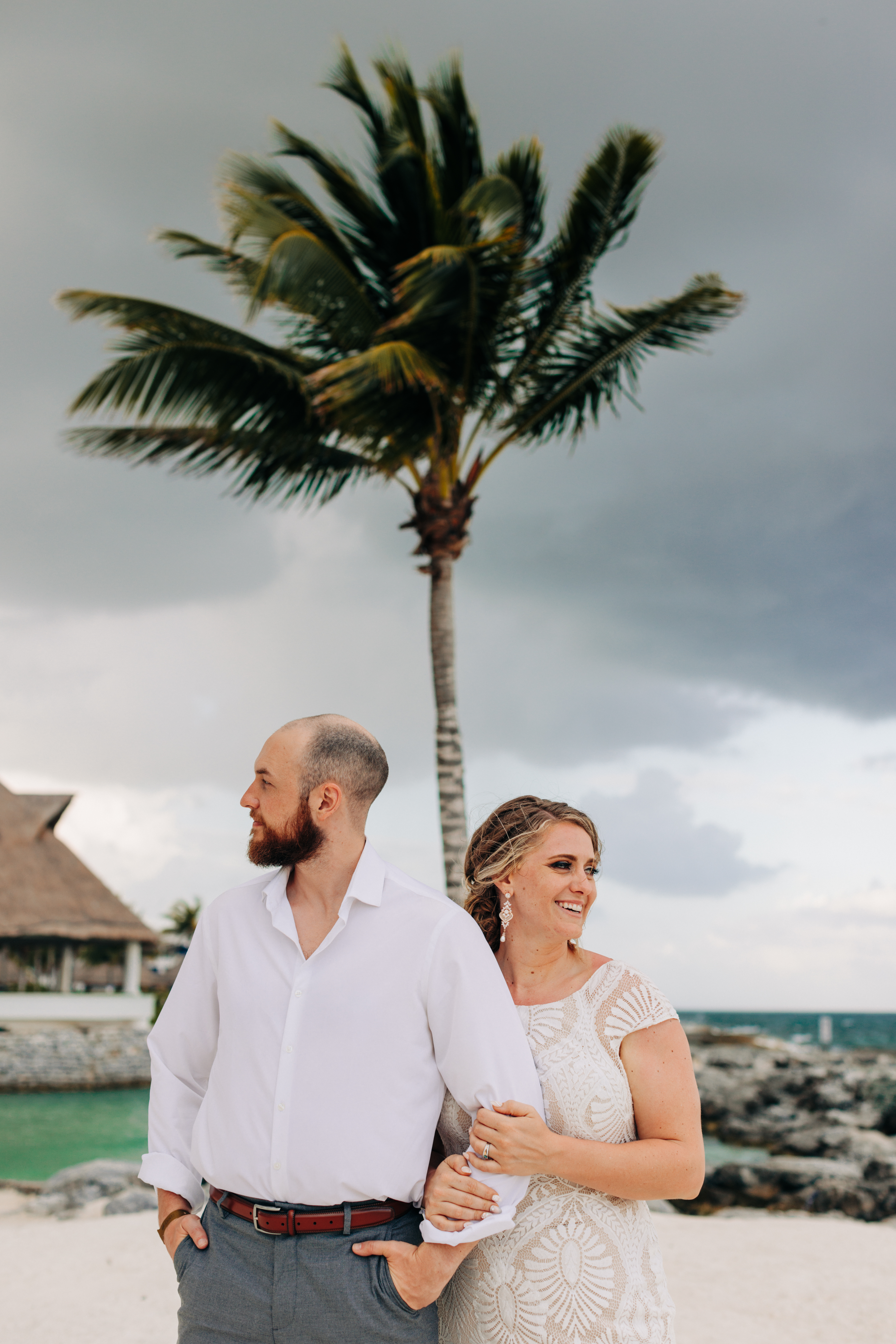 Bride holds onto grooms arm while they look in opposite directions with a palm tree between them